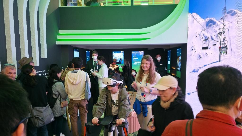 Obiskovalci na slovenskem paviljonu ob otvoritvi, ena obiskovalka sedi na kolesu, na glavi ima virtualna očala